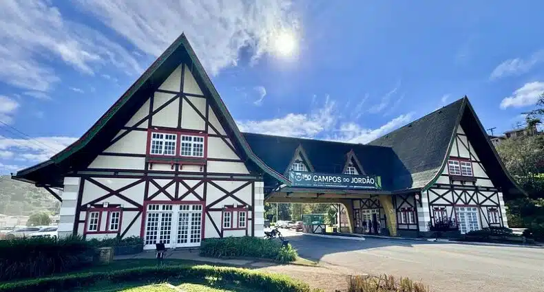 A imagem retrata a entrada da cidade de Campos do Jordão, em São Paulo. No centro, entre dois edifícios de arquitetura alpina, há um telhado com uma placa que diz o nome da cidade. O céu está ensolarado e com poucas nuvens.