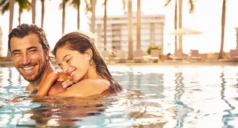 Imagem de um casal dentro da piscina do Four Seasons Hotel Miami durante o dia, com uma mulher do lado direito abraçada com um homem a frente da imagem. Representa hotéis românticos em Miami.