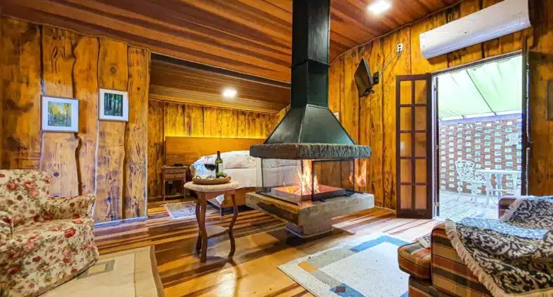 Quarto do Verde que te quero Ver-te - Chalés. Uma cama de casal está ao fundo, de frente para uma grande lareira no meio do ambiente. Ao lado esquerdo da lareira há uma poltrona e ao lado direito uma varanda. De frente para ela, no canto direito, está outra poltrona. A imagem ilustra o post de chalés em Visconde de Mauá.