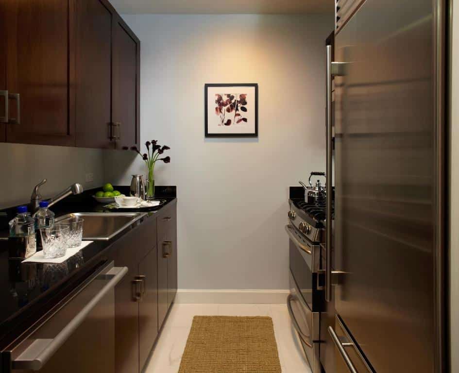 Cozinha compacta do Trump International hotel com armários de madeira escura, bancada de granito preto, e eletrodomésticos de aço inoxidável, incluindo geladeira e fogão. Há uma pequena obra de arte na parede. Este é um dos hotéis em Nova York.