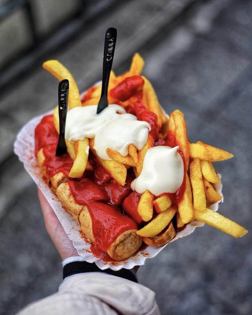 Imagem de uma pessoa segurando um prato com batatas fritas, salsicha e molhos em cima.