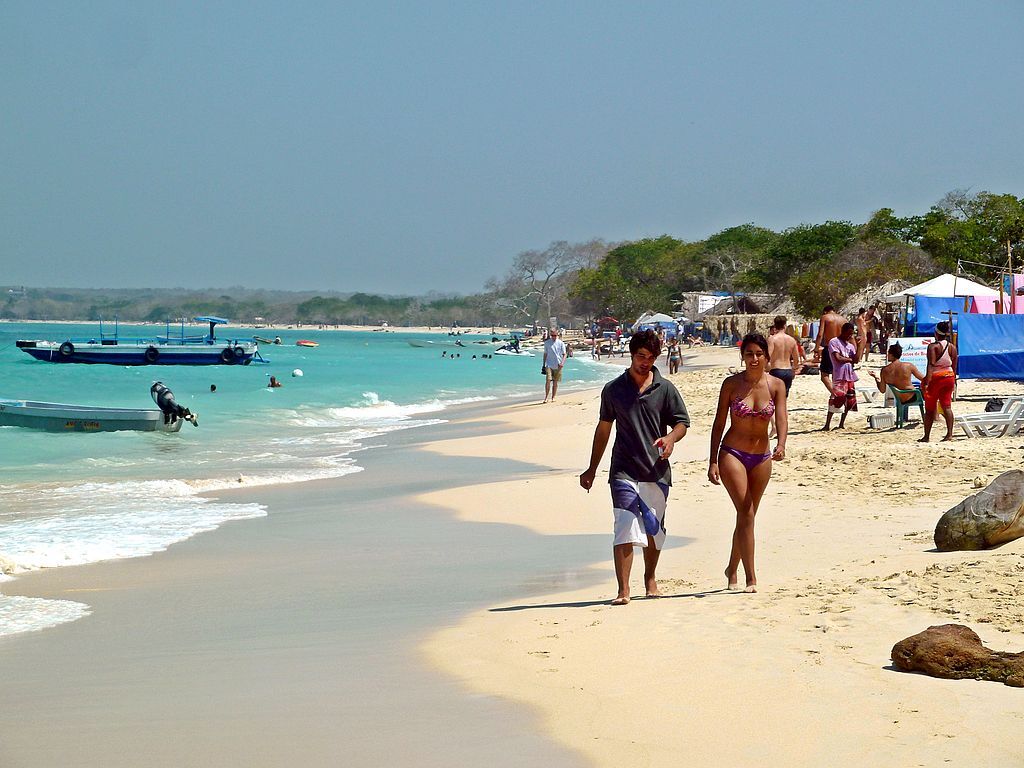 A imagem mostra a Playa Blanca, em Cartagena, movimentada com areia clara e mar azul-turquesa. Várias pessoas estão aproveitando o dia ensolarado, algumas nadando no mar e outras caminhando ou descansando na areia. Na frente, um casal caminha pela beira da praia, com barcos ancorados próximos à água. À direita, há cabanas e tendas coloridas, além de cadeiras de praia onde as pessoas relaxam. Ao fundo, a vegetação verde densa adiciona um toque natural ao cenário. - Foto: David Shankbone via Wikimedia Commons