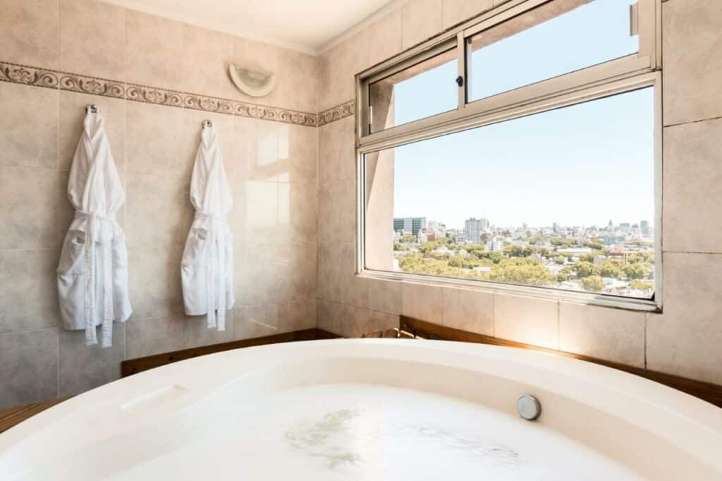 Uma banheira de hidromassagem do Days Inn by Wyndham Montevideo, com dois roupões pendurados no fundo e a janela. Imagem para ilustrar post sobre hotéis baratos em Montevidéu.