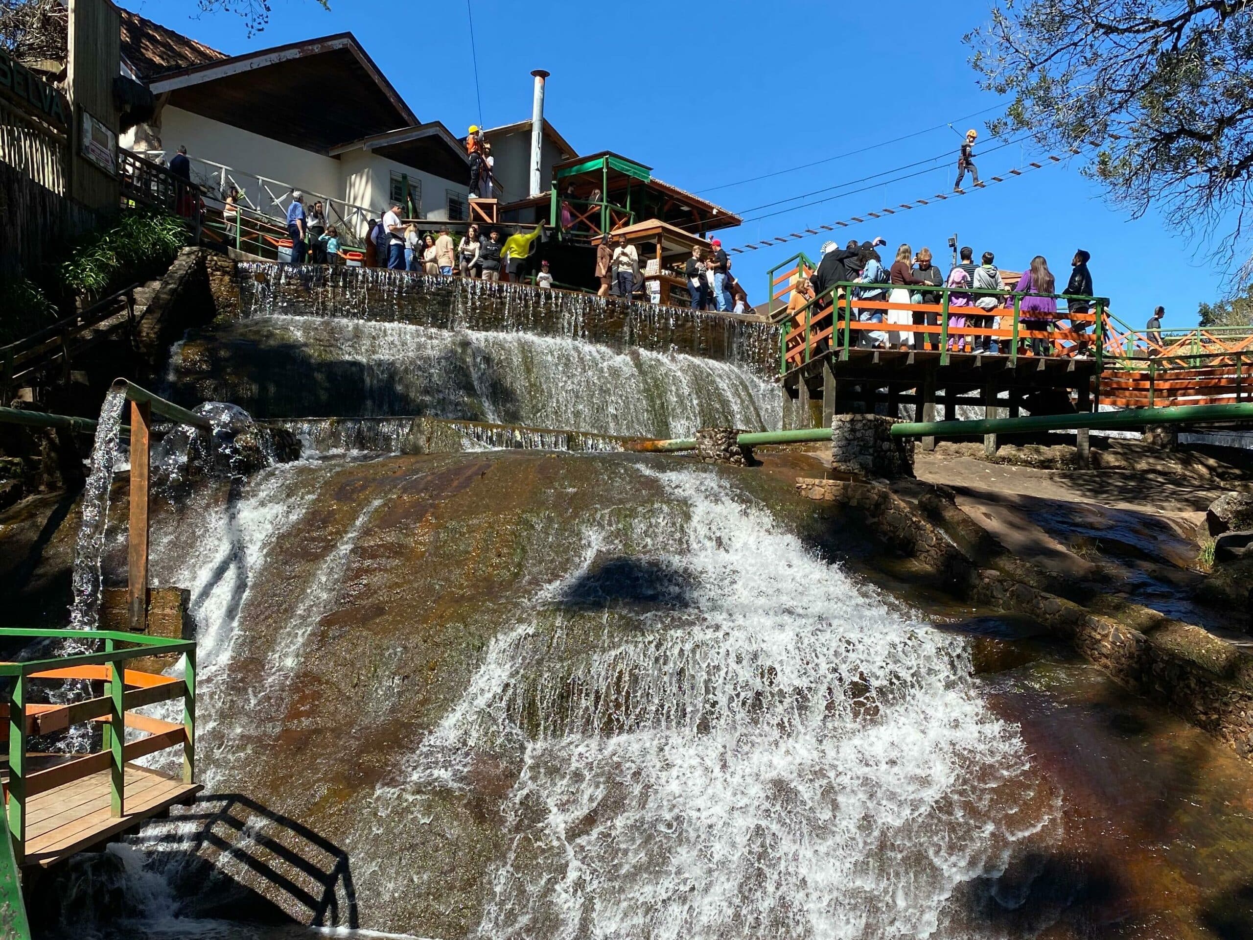 A imagem mostra uma cachoeira da Ducha de Prata em Campos do Jordão. Há pontes sobre a ducha onde as pessoas caminham. No topo se vê uma casa e uma ponte de tirolesa. O céu está azul.
