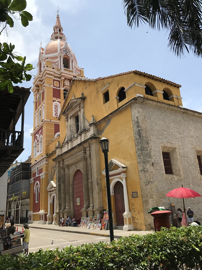 A imagem mostra a Igreja de Santo Domingo, durante o dia. A igreja apresenta uma fachada amarelo-ocre com detalhes arquitetônicos em pedra e uma grande porta vermelha de madeira. À esquerda, destaca-se uma torre alta e colorida, com tons de rosa, amarelo e branco, adornada com detalhes ornamentais. Em frente à igreja, há várias pinturas à venda, dispostas no chão ao lado de pessoas que descansam à sombra. À direita, uma pessoa segura um guarda-sol vermelho. Ao fundo, algumas árvores e um céu claro complementam a cena, que transmite a vivacidade e o encanto histórico do local. - Foto: Elias Rovielo via Flickr