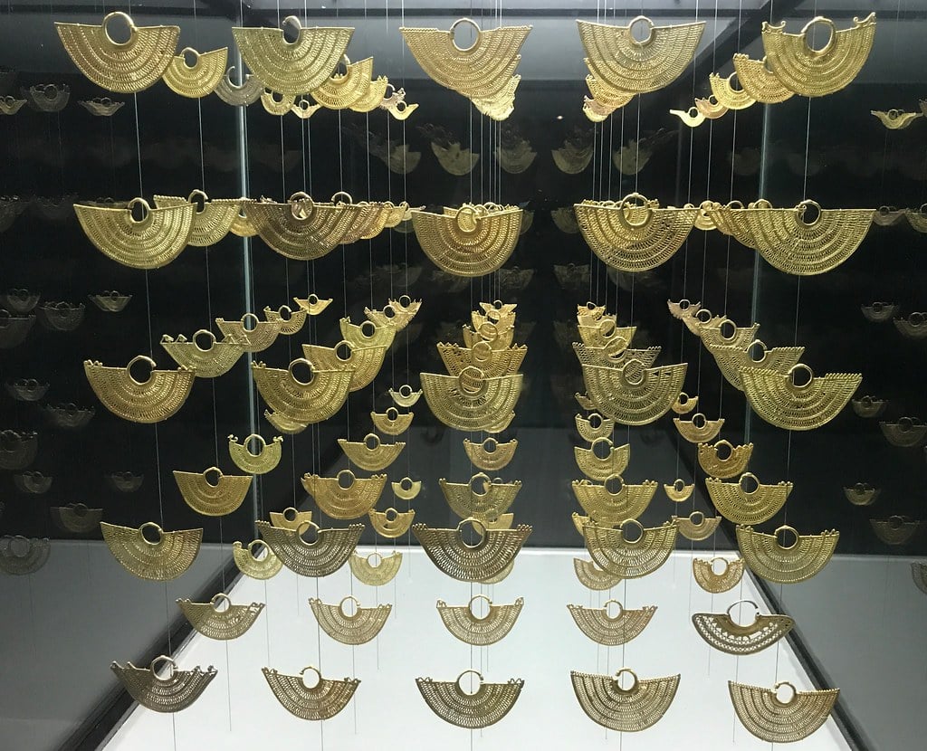 A imagem mostra uma exibição de peças de ouro em um museu, com várias joias penduradas em fios finos. As peças são em forma de leque semicircular, intrincadamente trabalhadas com padrões detalhados. Elas estão dispostas em várias fileiras e colunas, criando um efeito visual impressionante contra o fundo escuro. A disposição das peças permite que elas pareçam flutuar no ar, destacando a habilidade artesanal e a riqueza do material.