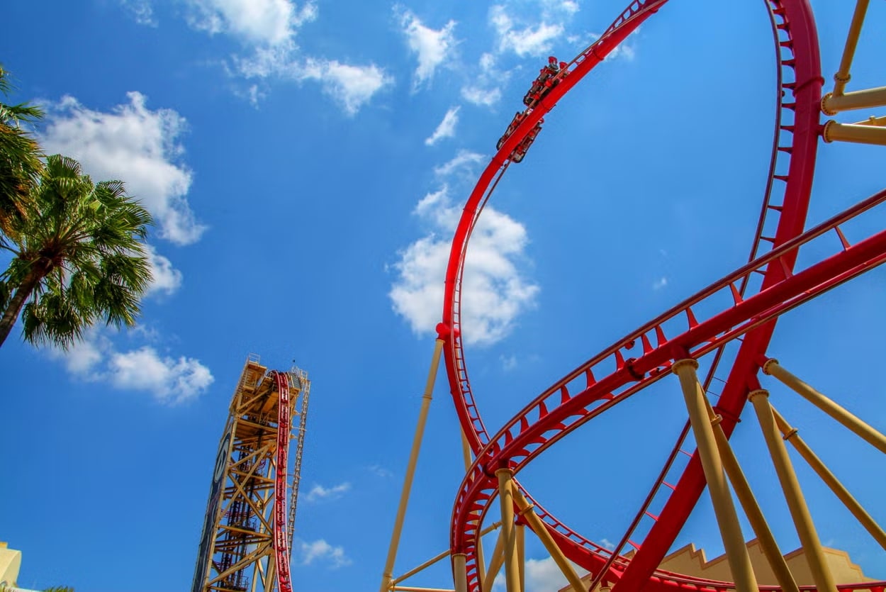 A imagem mostra uma impressionante montanha-russa com trilhos vermelhos em contraste com o céu azul claro e algumas nuvens na Universal Studios. A estrutura da montanha-russa inclui uma subida íngreme e uma grande curva em loop. Os visitantes são vistos no carrinho da montanha-russa enquanto experimentam a emoção da queda e da subida.
Ao lado esquerdo da imagem, há uma palmeira verde, adicionando um toque tropical ao ambiente. A estrutura da montanha-russa é sustentada por pilares amarelos, destacando-se ainda mais contra o céu azul.