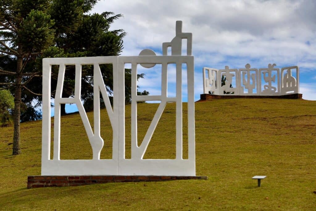 Duas esculturas brancas sobre um gramado verde. A que está na frente representa uma lua em uma janela. Há mais uma escultura no fundo da foto, em formato diferente. Na direita atrás da primeira escultura, há uma árvore. O céu está claro e com algumas nuvens.
