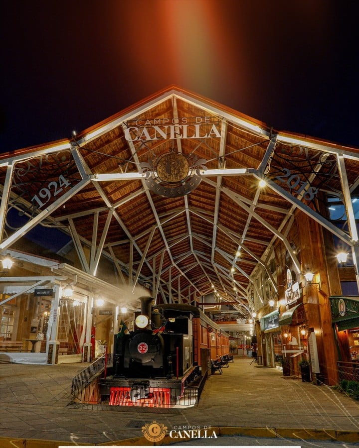 A imagem mostra a Estação Campos de Canella, um dos pontos turísticos de Canela, Rio Grande do Sul, durante a noite. A estrutura do telhado é composta por vigas de madeira e metal, com uma arquitetura que remete às estações ferroviárias antigas. No centro, há uma placa com os dizeres "Campos de Canella" e o ano de "1924" de um lado e "2018" do outro, indicando a preservação e revitalização do local. Em destaque, está uma antiga locomotiva a vapor, decorada com bandeiras do Brasil, sob a cobertura da estação. O ambiente é iluminado por luzes suaves, criando uma atmosfera nostálgica e aconchegante. Ao redor, vêem-se lojas e estabelecimentos que compõem o complexo da estação, tornando o local um atrativo turístico que combina história e modernidade.
