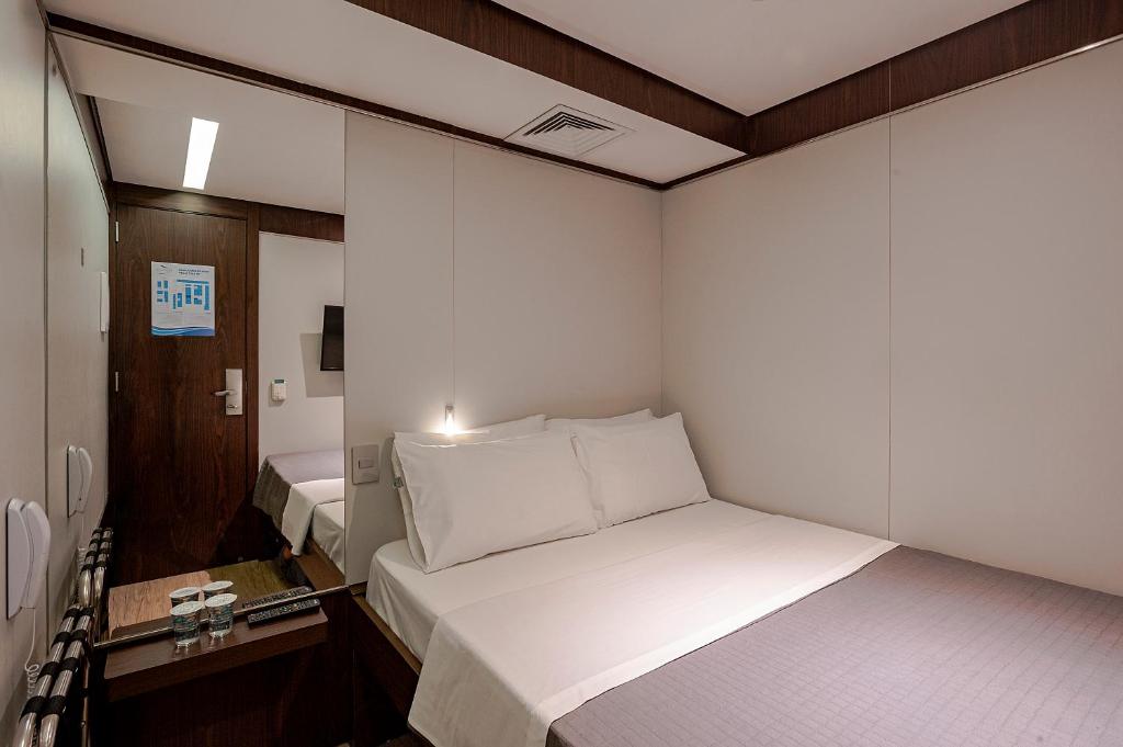Um pequeno quarto no FAST SLEEP Guarulhos by Slaviero Hotéis com uma cama de casal, uma mesa de cabeceira com garrafas de água e um espelho ao lado de uma porta de madeira, para representar hotéis perto do Aeroporto de Guarulhos.