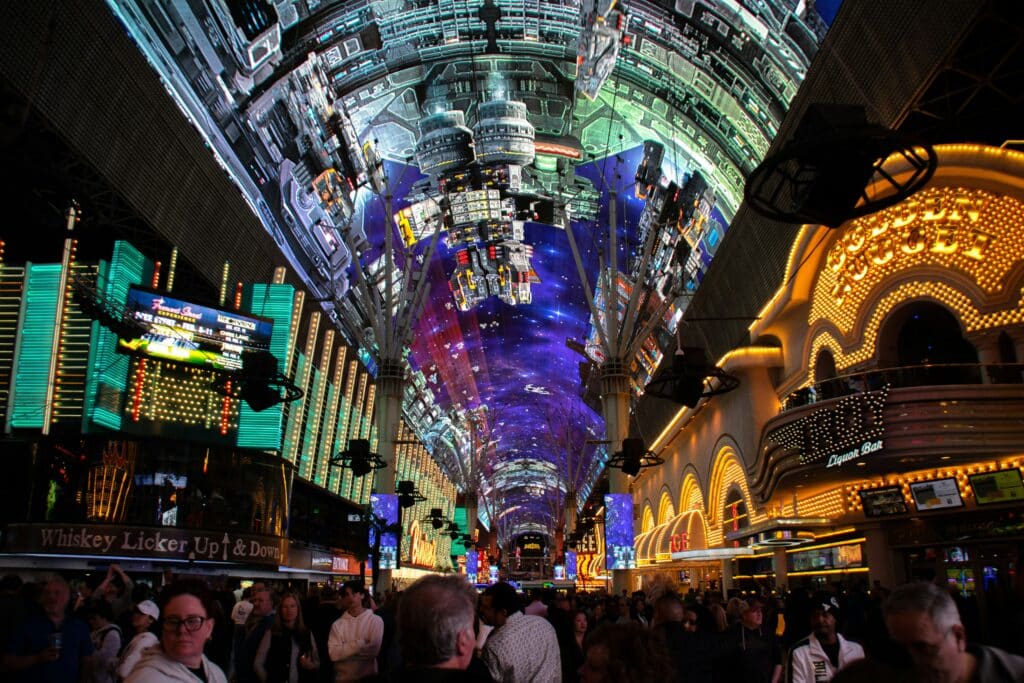 Teto de LED na Fremont Street. Ele se estende pela rua inteira e é todo colorido e iluminado, Diversas pessoas caminham abaixo dele. Do lado delas estão vários estabelecimentos, também cheios de luzes. A foto ilustra o post de onde ficar em Las Vegas.