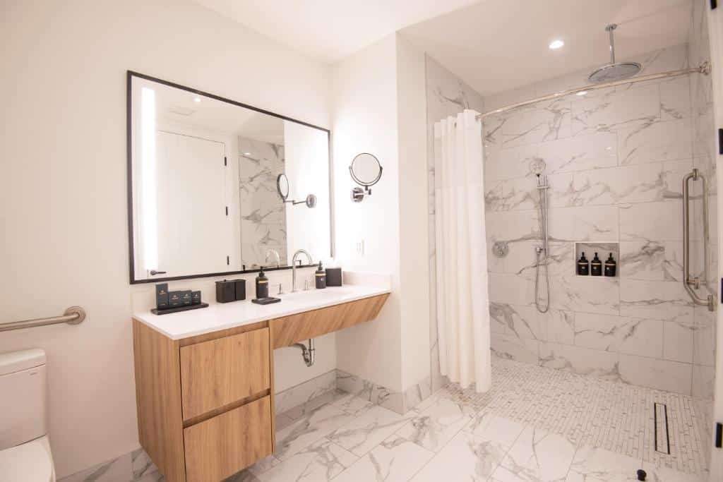 Banheiro do Goldwynn Resort & Residences que oferece recurso de acessibilidade para pessoas com mobilidade reduzida. Na parte esquerda tem um vaso sanitário com uma barra atrás, do lado tem um armário de pia para lavar as mãos, em cima na parede um espelho e ao lado um espelho pequeno redondo. Já ao fundo da imagem tem a parte do chuveiro sem box, apenas com uma cortina branca. Tem uma barra na vertical e um chuveiro. Imagem ilustrando post Hotéis nas Bahamas.