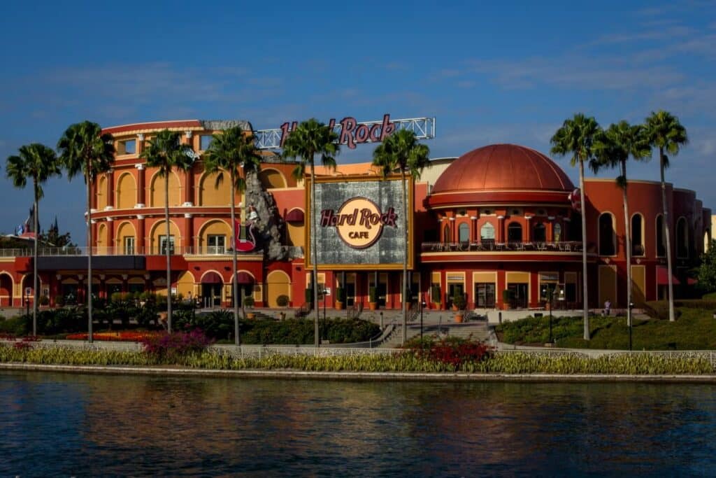 A imagem mostra a fachada do Hard Rock Cafe em Orlando, Flórida. O edifício é bastante imponente e colorido, com uma arquitetura que mistura elementos modernos e clássicos. As cores predominantes são o laranja e o vermelho, criando um visual vibrante e convidativo.
Na frente do prédio, há várias palmeiras altas, que adicionam um toque tropical ao ambiente. O logotipo do Hard Rock Cafe é claramente visível em um grande painel, que está centralizado na parte frontal da construção. Além disso, há uma grande guitarra decorativa, característica das decorações temáticas do Hard Rock Cafe, fixada na lateral do edifício.
O edifício possui várias janelas e sacadas, e o design geral apresenta um equilíbrio entre linhas retas e curvas.