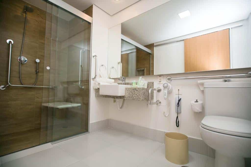 Banheiro com acessibilidade do Holiday Inn Parque Anhembi, an IHG Hotel. Um vaso sanitário do lado direito com barras de apoio e uma pia. Do lado esquerdo um box com barras de apoio. Em cima do vaso um espelho.