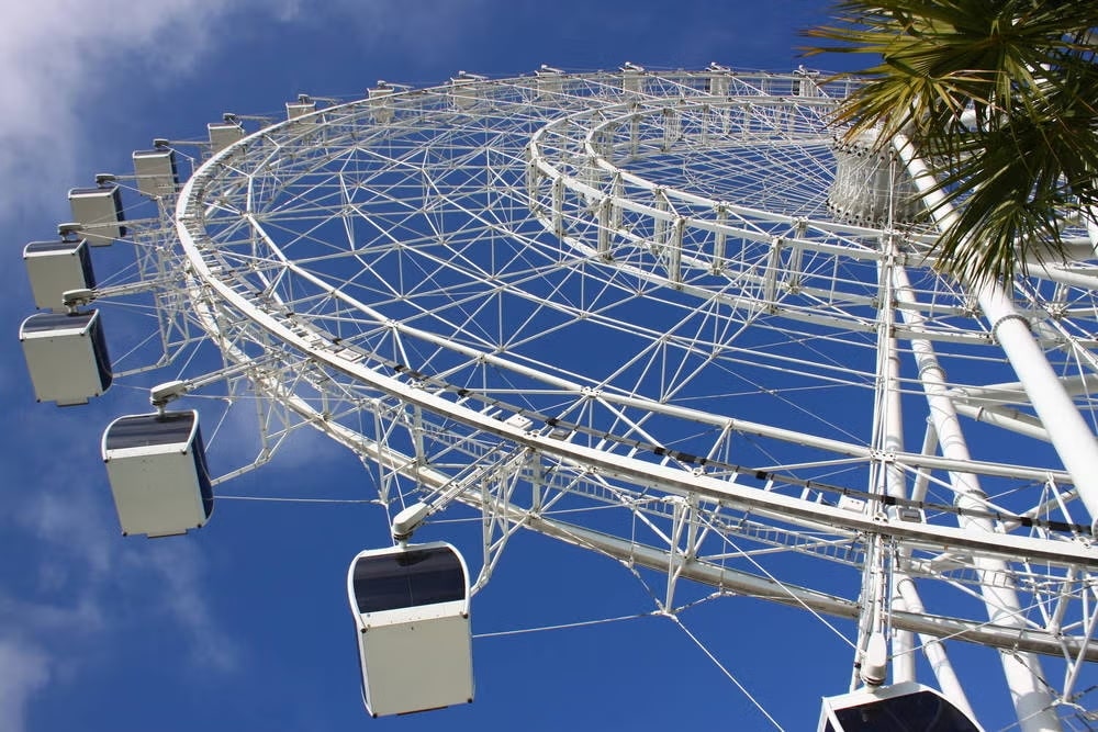 A imagem mostra a roda-gigante Icon Orlando 360, também conhecida como The Wheel, localizada em Orlando, Flórida. A foto foi tirada de um ângulo baixo, olhando para cima, destacando a imensa estrutura da roda contra um céu azul claro.
A estrutura é branca e composta de uma complexa rede de suportes e cabos, formando a roda gigante. As cabines são fechadas, com janelas de vidro, permitindo uma vista panorâmica da cidade para os passageiros. Algumas palmeiras são visíveis na parte direita da imagem, adicionando um elemento tropical ao cenário.