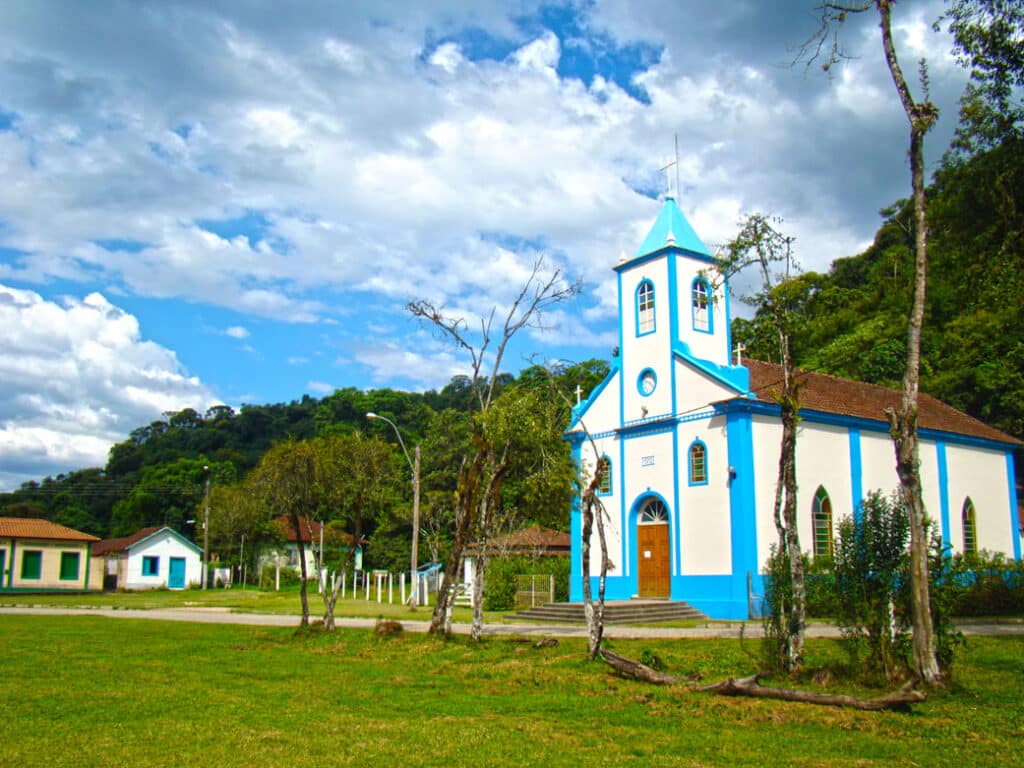 Igreja de São Sebastião na vila de Mauá com paredes brancas e azuis e bastante árvores verdes em volta. Perto da igreja também tem algumas casas. Imagem ilustrando post O que fazer em Visconde de Mauá.