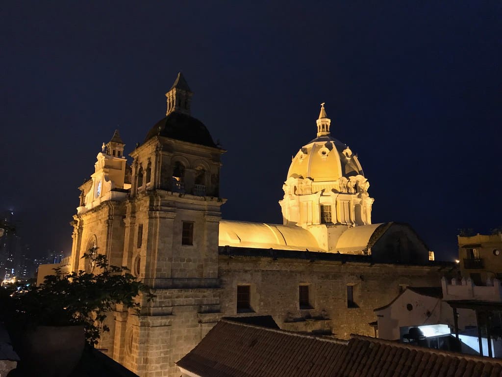 A imagem mostra a Igreja de San Pedro Claver, em Cartagena, iluminada à noite. A igreja possui uma arquitetura colonial com duas torres com campanários e uma grande cúpula central, todas destacadas por uma iluminação amarela que realça os detalhes da construção. O céu noturno escuro proporciona um contraste marcante com a iluminação da igreja, criando uma cena impressionante. À esquerda, é possível ver algumas luzes da cidade ao fundo, adicionando um toque urbano à imagem.