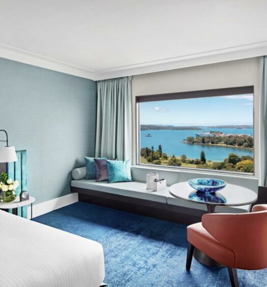 Quarto do InterContinental Sydney, an IHG Hotel para ilustrar post sobre hotéis em Sydney. Uma cama de casal do lado esquerdo com uma luminária. Do lado direito uma poltrona, uma mesa e um abajur. No fundo um sofá embaixo da janela com vista do mar.