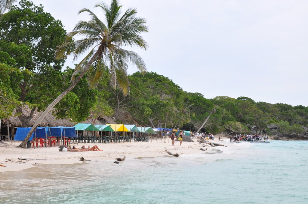 A imagem mostra uma praia tropical chamada Playa Blanca, em Cartagena, com areia branca e fina e águas cristalinas. À esquerda, há uma palmeira inclinada e uma fileira de cabanas e tendas coloridas (azuis, verdes e amarelas) com mesas e cadeiras vermelhas debaixo delas. Pessoas relaxam na areia e caminham pela praia. Ao fundo, uma vegetação densa e verdejante, criando uma sensação de natureza preservada. À direita, um grupo de pessoas se reúne perto de um barco ancorado na água. - Foto: Jorge Láscar via Flickr