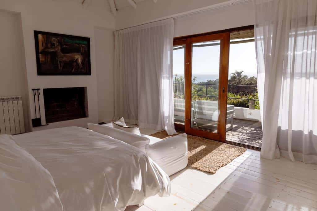Quarto bem iluminado na La Posada del Faro com roupa de cama branca, poltronas, lareira, portas de vidro e vista para um pátio e vegetação externa.