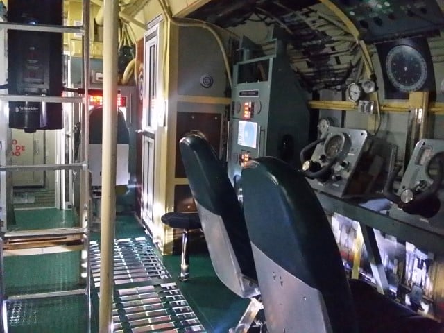 A imagem mostra o interior de um submarino em exposição no Museu Naval do Caribe. O espaço é apertado e repleto de equipamentos e painéis de controle. Há duas cadeiras pretas posicionadas em frente a um painel com vários mostradores, botões e medidores. Uma escada metálica vertical leva a um nível superior. A iluminação é suave, destacando as texturas e os detalhes dos instrumentos.