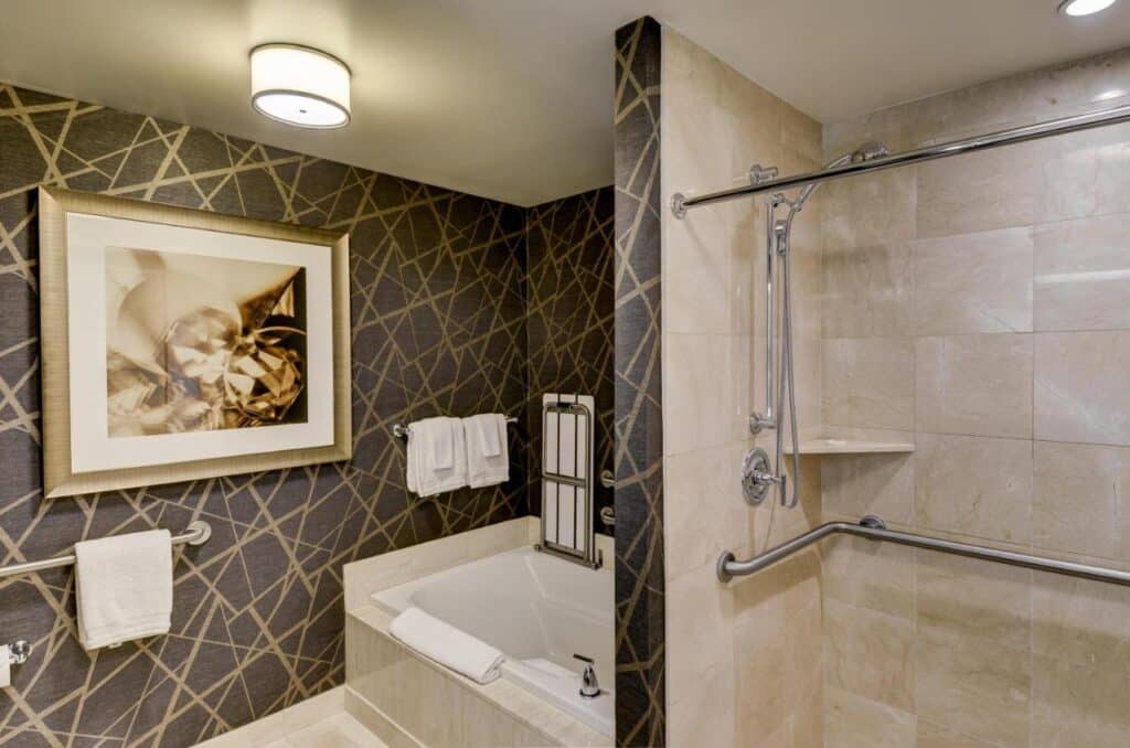 Banheiro acessível do Mandalay Bay. Do lado direito está a área do chuveiro, com uma grande barra de apoio na parede inteira. Do lado esquerdo está uma banheira, um quadro na parede e outra barra de apoio abaixo dele.
