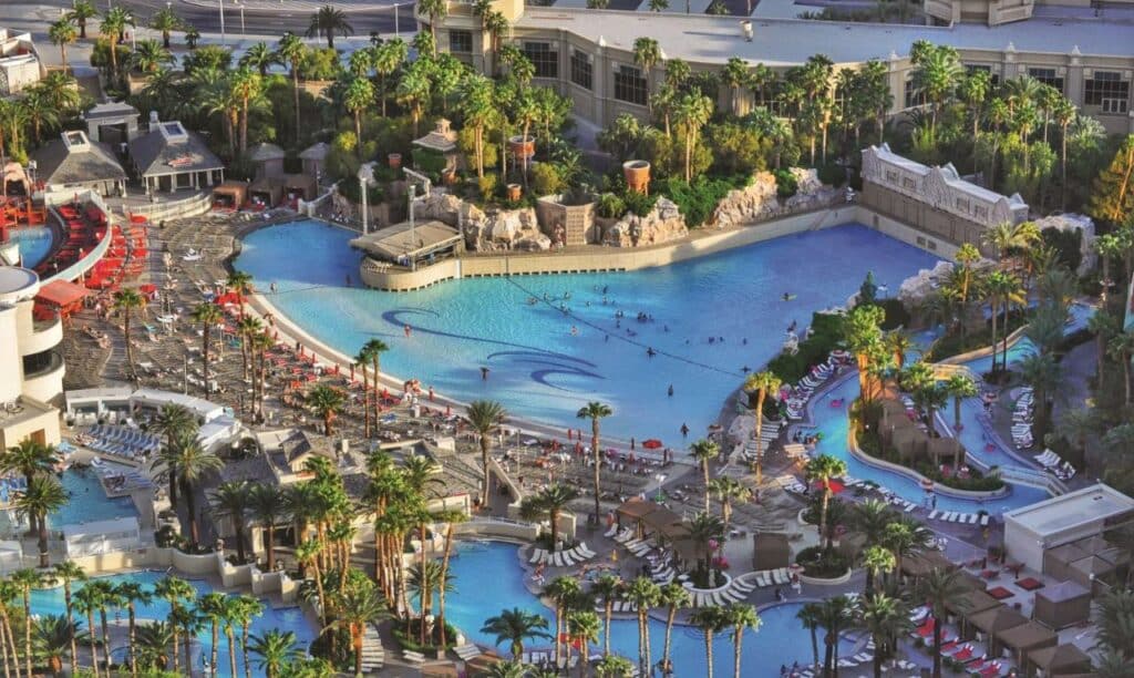 Vista de cima de uma das piscinas do Mandalay Bay. Ela está do lado direito e tem elevações para formar ondas. Ao redor estão vários outras piscinas menores, árvores, cabamas e pessoas passando.