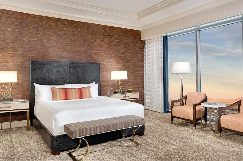 Quarto no Mandalay Bay com uma cama de casal com recamier no centro. Dos seus dois lados estao mesinhas com abajur. Do lado esquerdo está uma janela bem grande com vista para a cidade, além de duas poltronas e uma mesa entre elas.