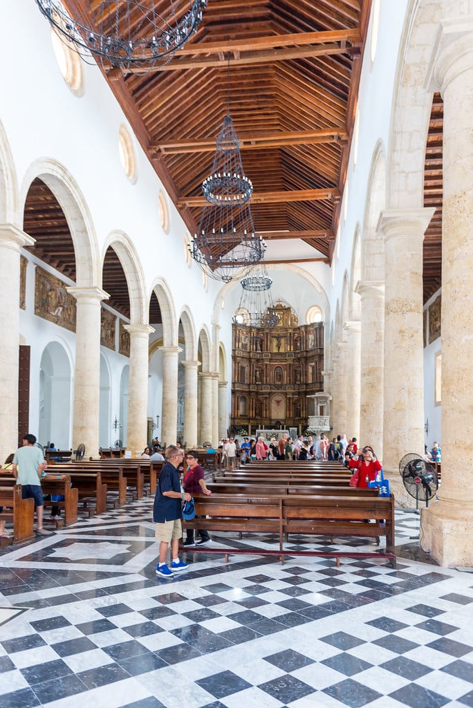 A imagem mostra o interior da Catedral Santa Catarina de Alexandria, em Cartagena. O piso é de mármore em padrão xadrez preto e branco, e a nave é ladeada por grandes colunas de pedra que sustentam arcos altos. O teto de madeira é adornado com lustres grandes de ferro forjado. No fundo, o altar principal é detalhadamente esculpido e ricamente decorado. Há várias pessoas dentro da igreja, algumas sentadas nos bancos de madeira e outras caminhando ou admirando a arquitetura. A luz natural entra através das janelas redondas altas, iluminando suavemente o ambiente. - Foto: MikePScott via Flickr