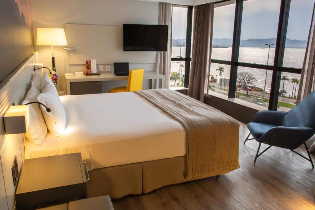Quarto do Novotel Florianopolis. Do lado esquerdo uma cama de casal com uma luminária de cada lado. No fundo uma mesa de trabalho com TV. Do lado direito uma poltrona e uma parede de vidro com vista do mar.