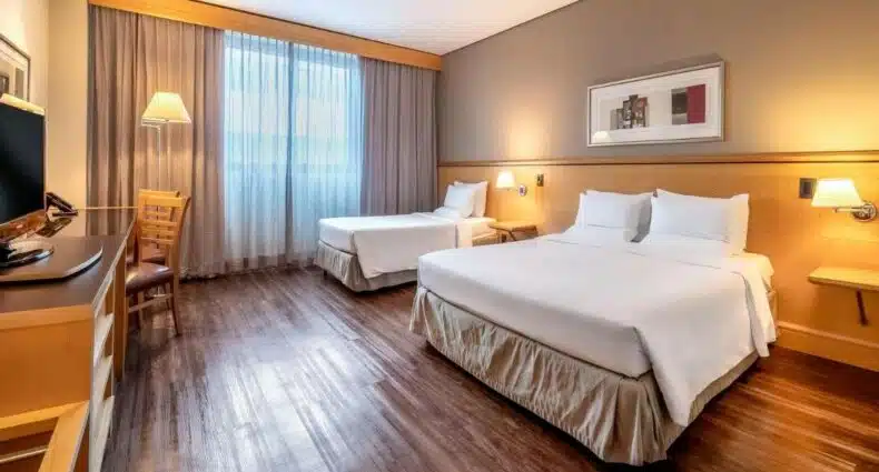 Quarto do Novotel Sao Paulo Center Norte, para ilustrar post sobre hotéis perto do Expo Center Norte. Do lado direito uma cama de casal e uma cama de solteiro, entre elas um abajur e uma prateleira. De frente uma raque, uma mesa de trabalho e uma TV. No fundo a janela com cortina.