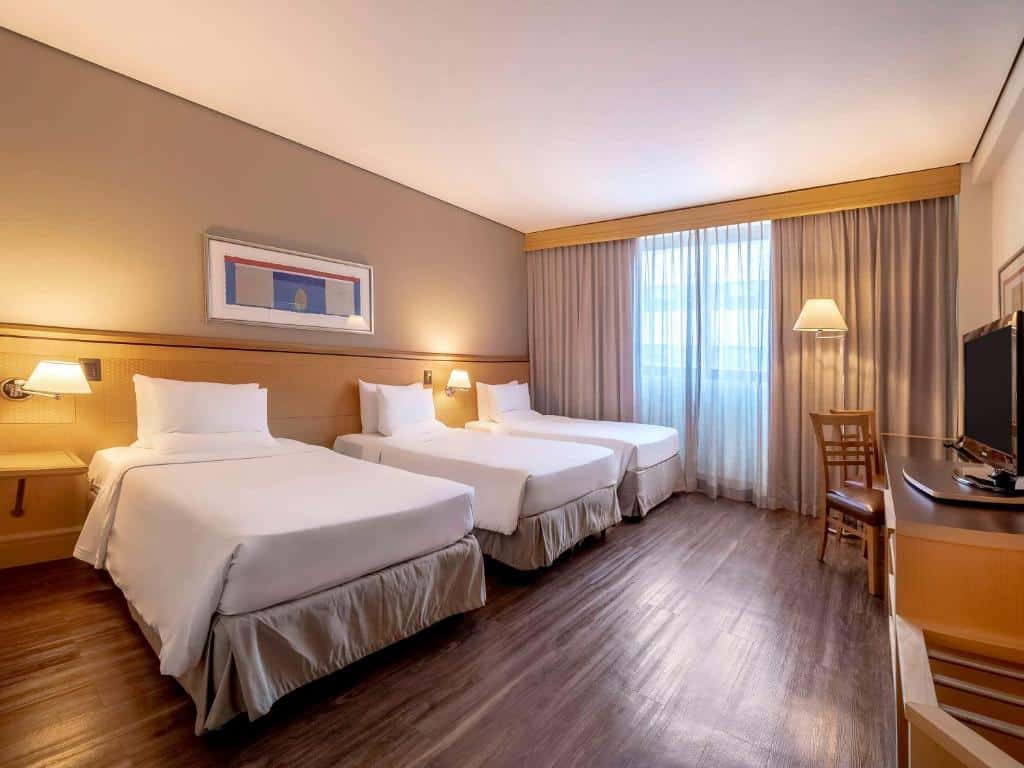 Quarto do Novotel Sao Paulo Center Norte. Três camas de solteiro do lado esquerdo, com dois abajures. Do lado direito uma mesa de trabalho, umta TV e um abajur. No fundo uma janela com cortina. Imagem para ilustrar post sobre hotéis perto do Expo Center Norte.