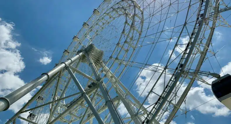 vista de baixo para cima da Orlando Eye uma das opções de o que fazer em Orlando, sendo uma roda gigante alta com estrutura em ferro e cabines grandes, o céu está azul com nuvens, sendo o clima ideal para visitar a atração e ter uma visão completa da cidade e arredores