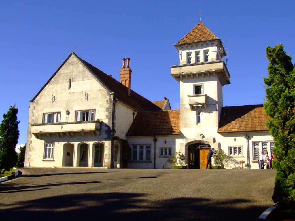 Imagem da fachada do Palácio Boa Vista em Campos do Jordão. É uma construção tradicional e requintada, com várias janelas e até mesmo uma torre. Há algumas pessoas em frente do palácio tirando foto. O céu está azul e sem nuvens. Há árvores ao redor do palácio. A imagem ilustra o post de o que fazer em Campos do Jordão.