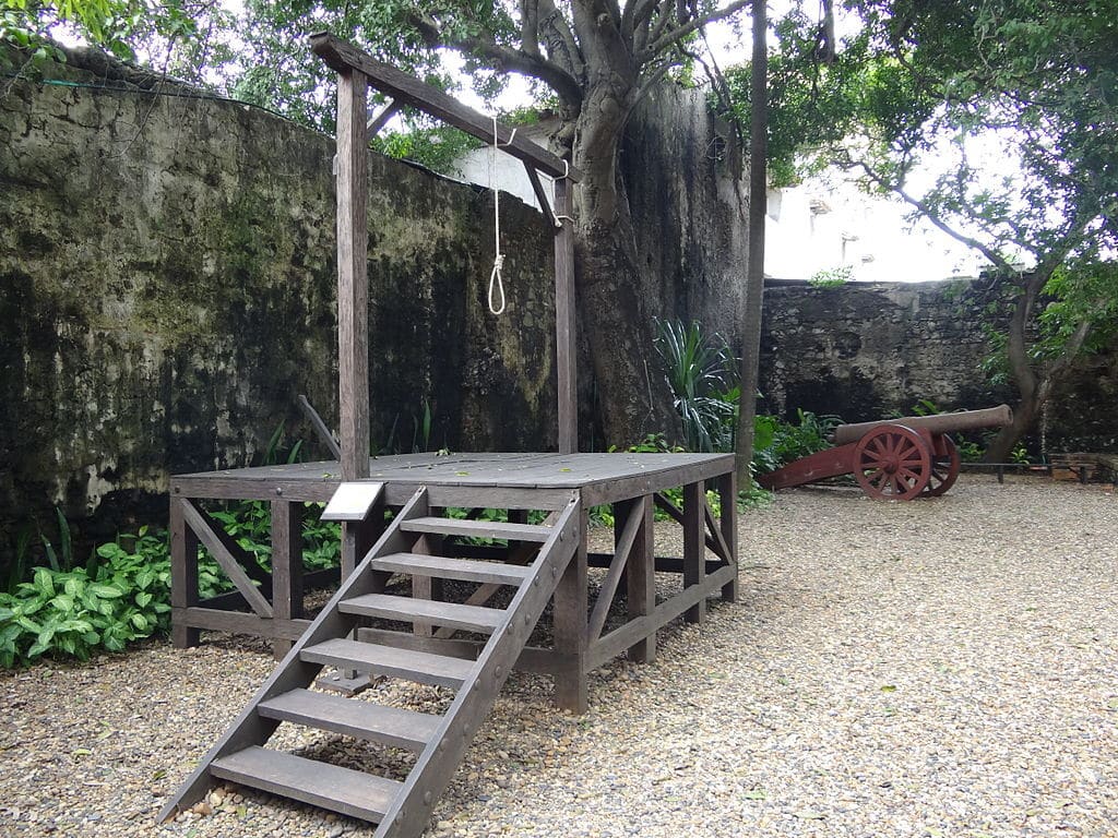 A imagem mostra um patíbulo de madeira, também conhecido como cadafalso, em um pátio de cascalho cercado por paredes de pedra envelhecida. O patíbulo possui uma plataforma elevada com escadas que levam ao topo, onde há uma estrutura com uma corda pendurada, indicando seu uso histórico para execuções. Ao fundo, à direita, há um antigo canhão vermelho apoiado no chão, apontando para cima, cercado por vegetação. - Foto: Kamilokardona via Wikimedia Commons