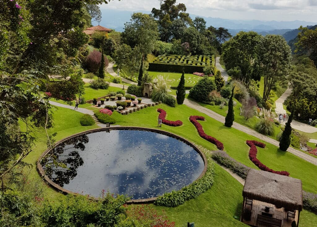 Vista aérea do Parque Amantikir em Campos do Jordão. Todo o espaço possui uma bela grama verde. No centro há um lago, e no canto inferior direito há um quiosque. Atrás do lado há um espaço com o que parecem ser mesas e cadeiras de arbusto, e ainda mais ao fundo há um labirinto. Há árvores por todo o local. A foto representa o post de o que fazer em Campos do Jordão.