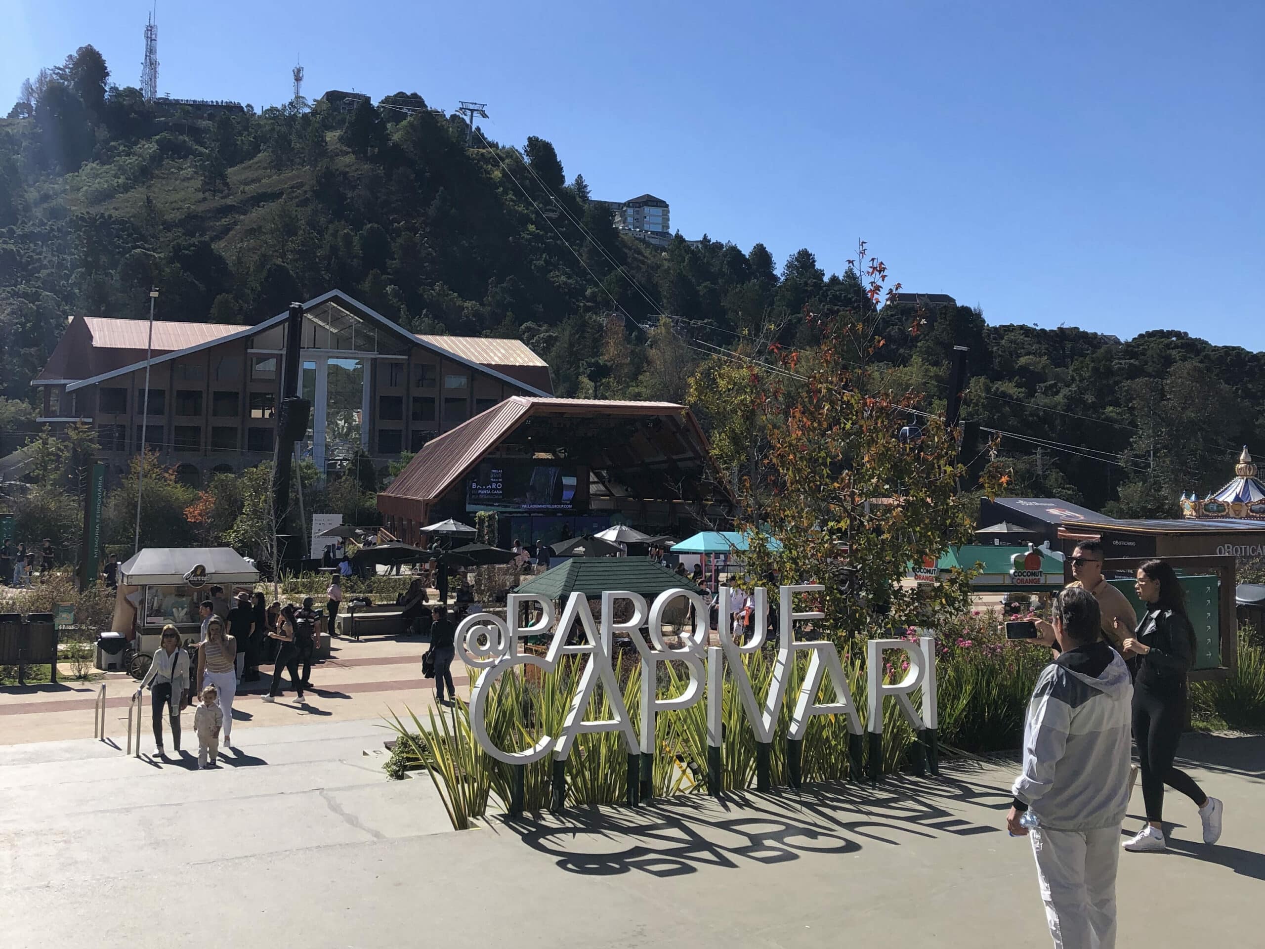Entrada do Parque Capivari, em Campos do Jordão. No calçadão do parque já vemos um letreiro no chão com o nome do parque entre alguns arbustos, ideal para tirar fotos. No fundo vemos um palco, barraquinhas de comida, pessoas caminhando e um monte por onde sobe um teleférico. Representa o post sobre o que fazer em Campos do Jordão.