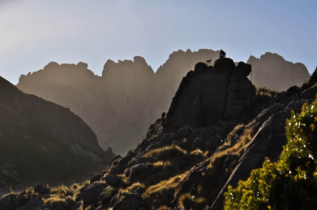 Parte do Parque Nacional do Itatiaia que mostra várias pedras com plantas em volta. Já em primeiro plano tem algumas pessoas escalando uma pedra da paisagem. 
