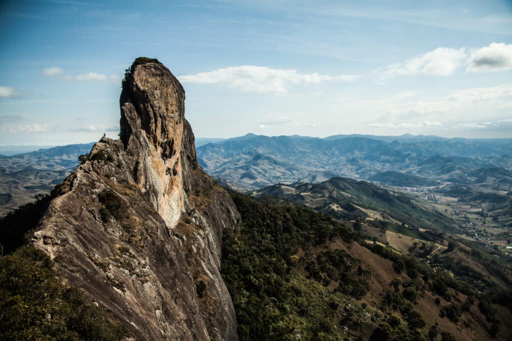 Vista aérea para a Pedra do Baú, uma formação rochosa que se alonga ao céu. Embaixo há montanhas que vão até o horizonte. O céu está claro e com algumas nuvens. A foto ilustra o post sobre o que fazer em Campos do Jordão.