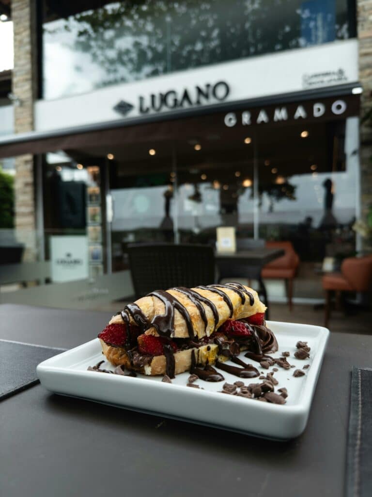 A imagem mostra uma sobremesa deliciosa na frente da loja Lugano, em Gramado. Em destaque no primeiro plano, há um prato retangular branco contendo uma sobremesa de croissant recheado com morangos frescos e coberto com calda de chocolate. Ao lado da sobremesa, há pedaços de chocolate espalhados pelo prato, realçando a apresentação.
Ao fundo, a fachada da loja Lugano é visível, com o nome "Lugano" em letras grandes e pretas sobre uma faixa branca, e "Gramado" escrito logo abaixo. A entrada da loja possui portas de vidro, através das quais se pode ver o interior iluminado e decorado de forma acolhedora. Há mesas e cadeiras na área externa, sugerindo um ambiente convidativo para apreciar doces e bebidas.