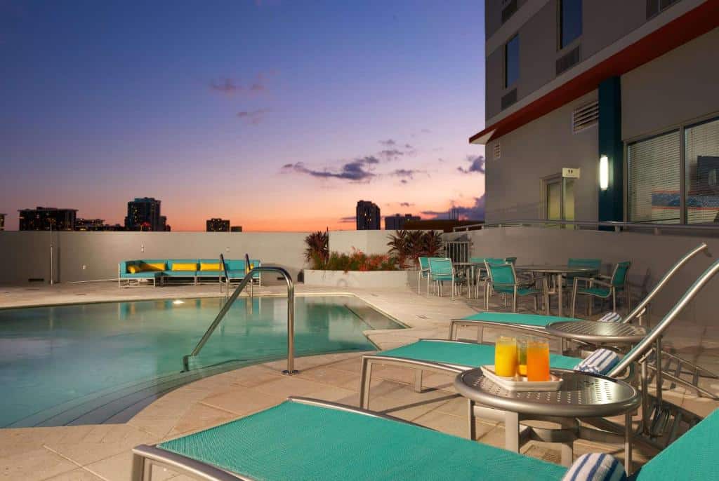 Área da piscina na cobertura do Hampton Inn & Suites by Hilton Miami Downtown/Brickell ao pôr do sol com espreguiçadeiras, mesas e bebidas. Horizonte da cidade visível à distância sob um céu colorido.
