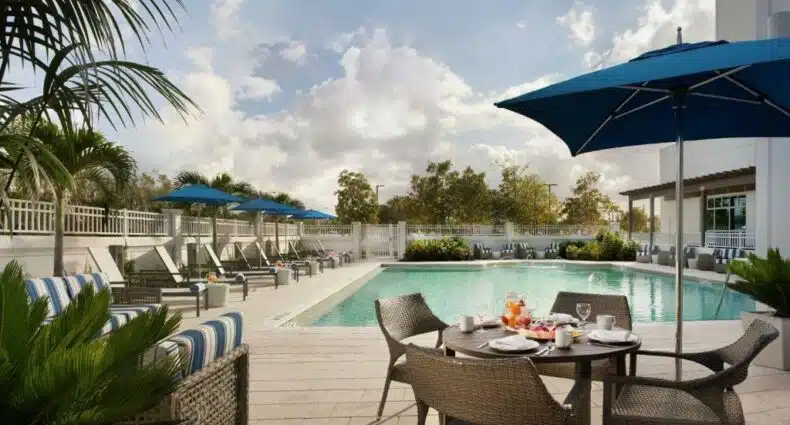 Área de lazer do Hilton Garden Inn Miami Dolphin Mall durante o dia com uma mesa com quatro cadeiras do lado esquerdo sofás e ao fundo no lado direito uma piscina quadrada.