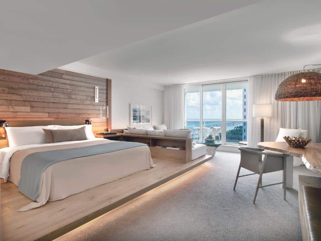 Quarto do 1 Hotel South Beach com cama king-size, parede com detalhes em madeira, escrivaninha e área de estar com vista para o mar através de grandes janelas. Representa onde ficar em Miami.