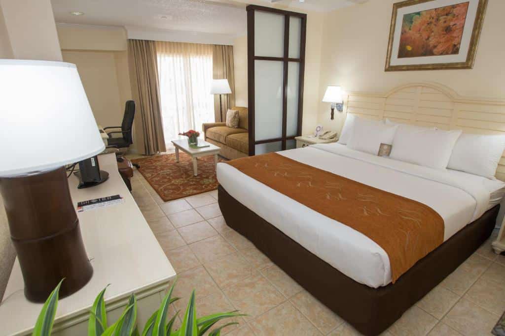 Quarto do Comfort Suites Paradise Island que mostra uma cama de casal com travesseiros e cobertas, do lado direito da cama uma mesa de cabeceira e em frente a cama tem uma cômoda com um abajur e alguns objetos. Já do lado direito do quarto ao fundo tem uma parte separada que parece uma sala com um sofá, um tapete no chão com uma mesinha de centro, uma cadeira e uma janela com cortinas.