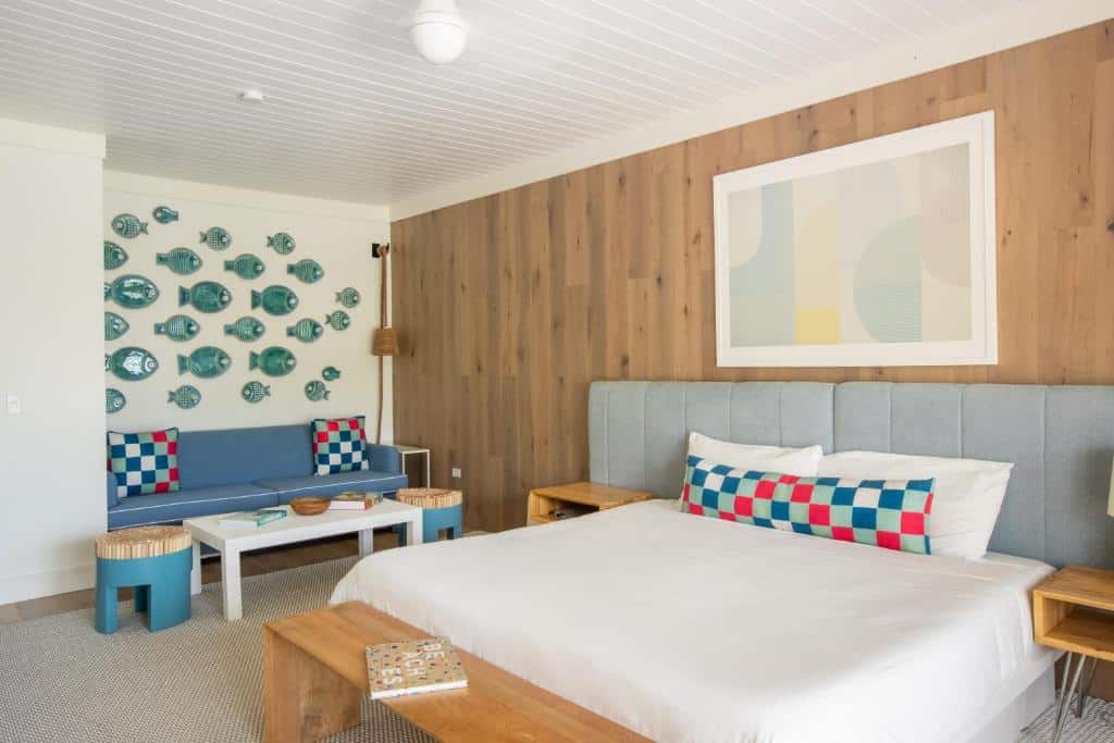 Quarto do Coral Sands Hotel que mostra uma cama de casal com travesseiros e coberta, de cada lado da cama tem uma mesa de cabeceira e nos pés da cama tem um banco de madeira. Do lado direito da cama ao fundo do quarto tem um sofá encostado na parede com duas almofadas uma mesinha retangular e dois bancos redondos.