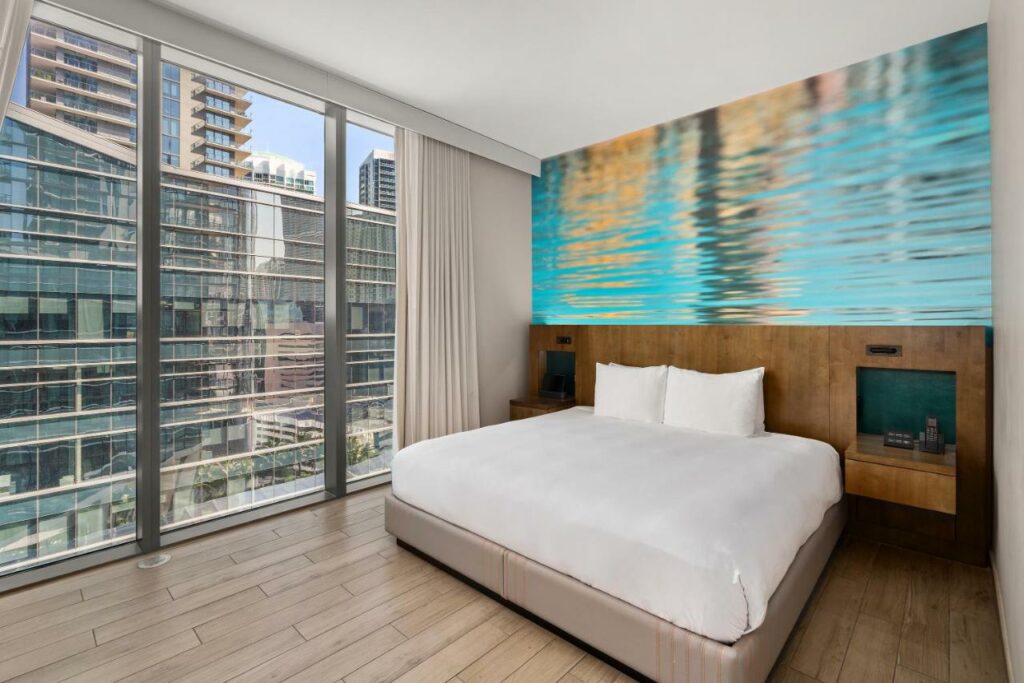 Quarto do EAST Miami moderno com uma cama grande, parede de vidro, piso de madeira e uma obra de arte colorida acima da cabeceira, com vista para os edifícios da cidade. Representa onde ficar em Miami.