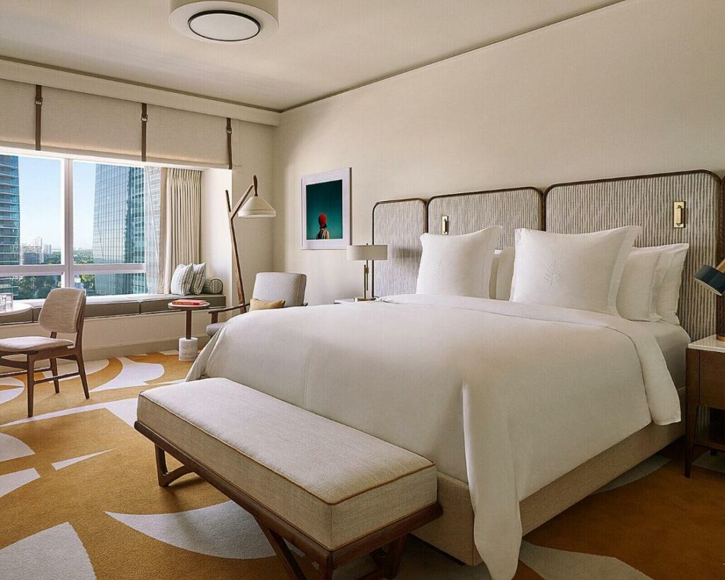 Quarto do Four Seasons Hotel Miami com cama de casal do lado direito da imagem, no pé da cama um banco estofado e do lado direito uma cômoda com luminária, ainda do lado esquerdo uma poltrona. Representa hotéis para Brasileiros em Miami.