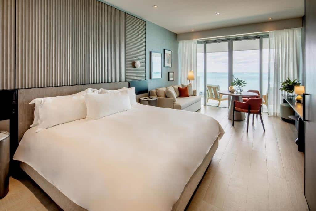 Quarto do Goldwynn Resort & Residences com uma grande cama de casal e de cada lado da cama tem uma mesinha redonda. Do lado esquerdo da cama tem uma parte estilo sala de estar com um sofá, uma mesa com duas cadeiras e uma cômoda. Do lado dessa área tem uma porta grande de vidro com cortinas que dá acesso a varanda do quarto com vista para a paisagem.