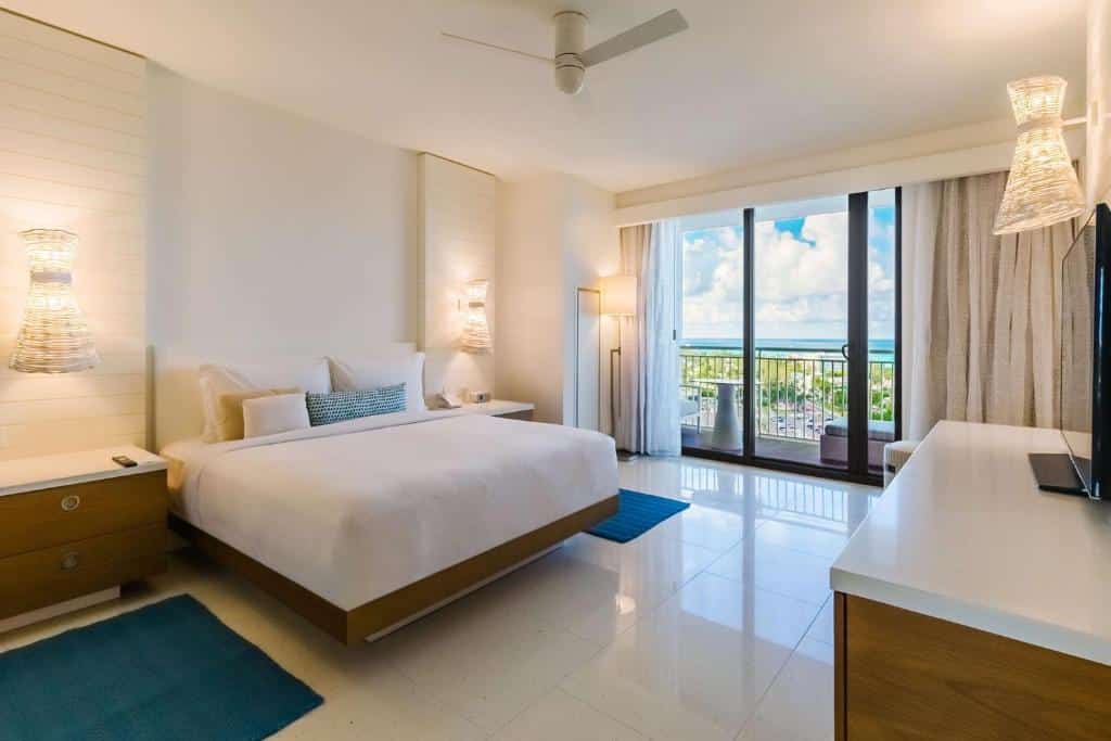 Quarto do Grand Hyatt Baha Mar que mostra uma cama de casal ao centro do quarto e de cada lado da cama tem uma mesa de cabeceira, uma luminária na parede e um tapete no chão. Em frente a cama tem um móvel de madeira com uma televisão em cima e do lado esquerdo do quarto tem uma porta grande de vidro com cortinas que dá acesso a varanda do quarto.