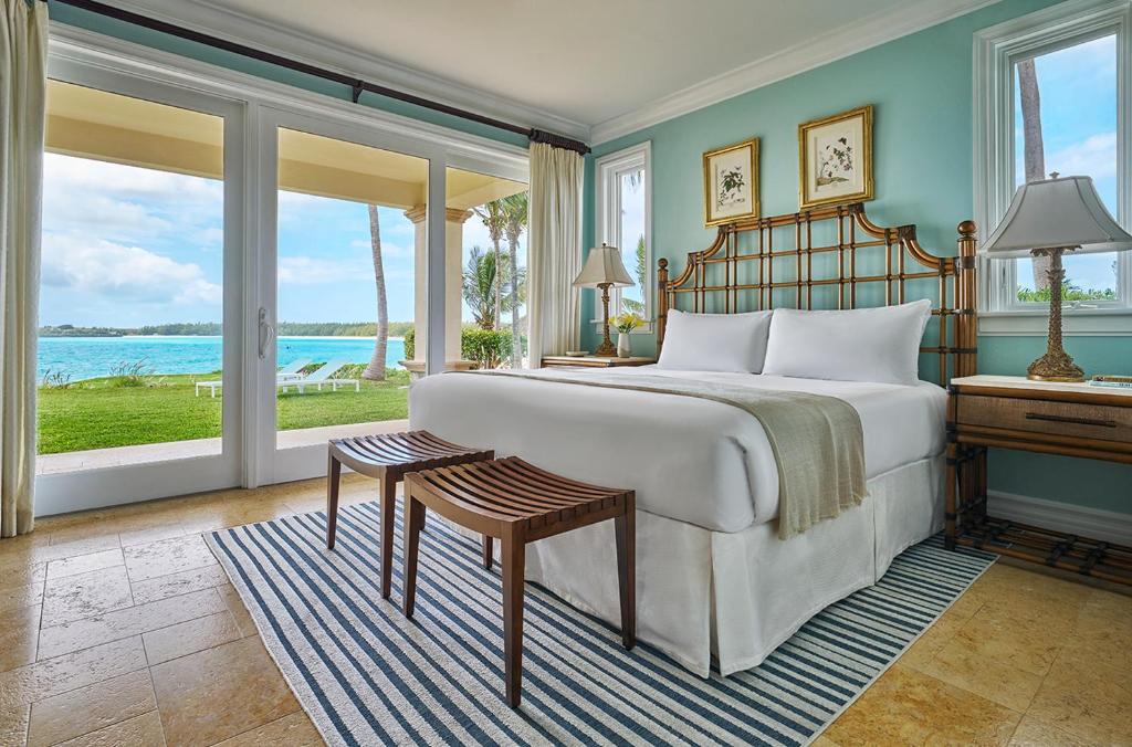 Quarto do Grand Isle Resort & Residences que mostra uma cama de casal, de cada lado da cama tem uma mesa de cabeceira com um abajur e nos pés da cama tem dois bancos de madeira. Do lado direito da cama tem uma grande porta de vidro com cortinas que dá acesso a uma área restrita com um gramado verde e vista para o mar.